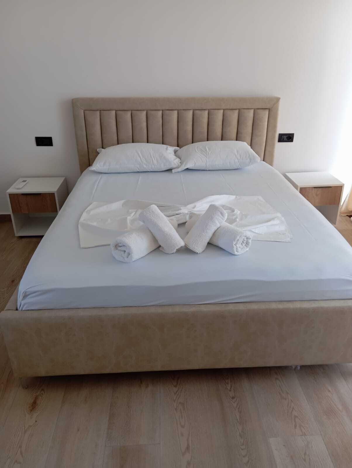 Una cama bien tendida con sábanas blancas y toallas enrolladas, un cabecero tapizado en beige y mesitas de noche de madera con un diseño sencillo.