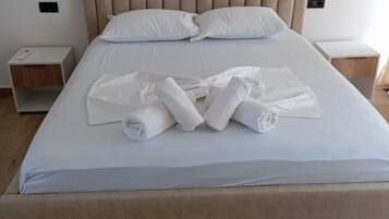 Una cama bien tendida con sábanas blancas y toallas enrolladas, un cabecero tapizado en beige y mesitas de noche de madera con un diseño sencillo.