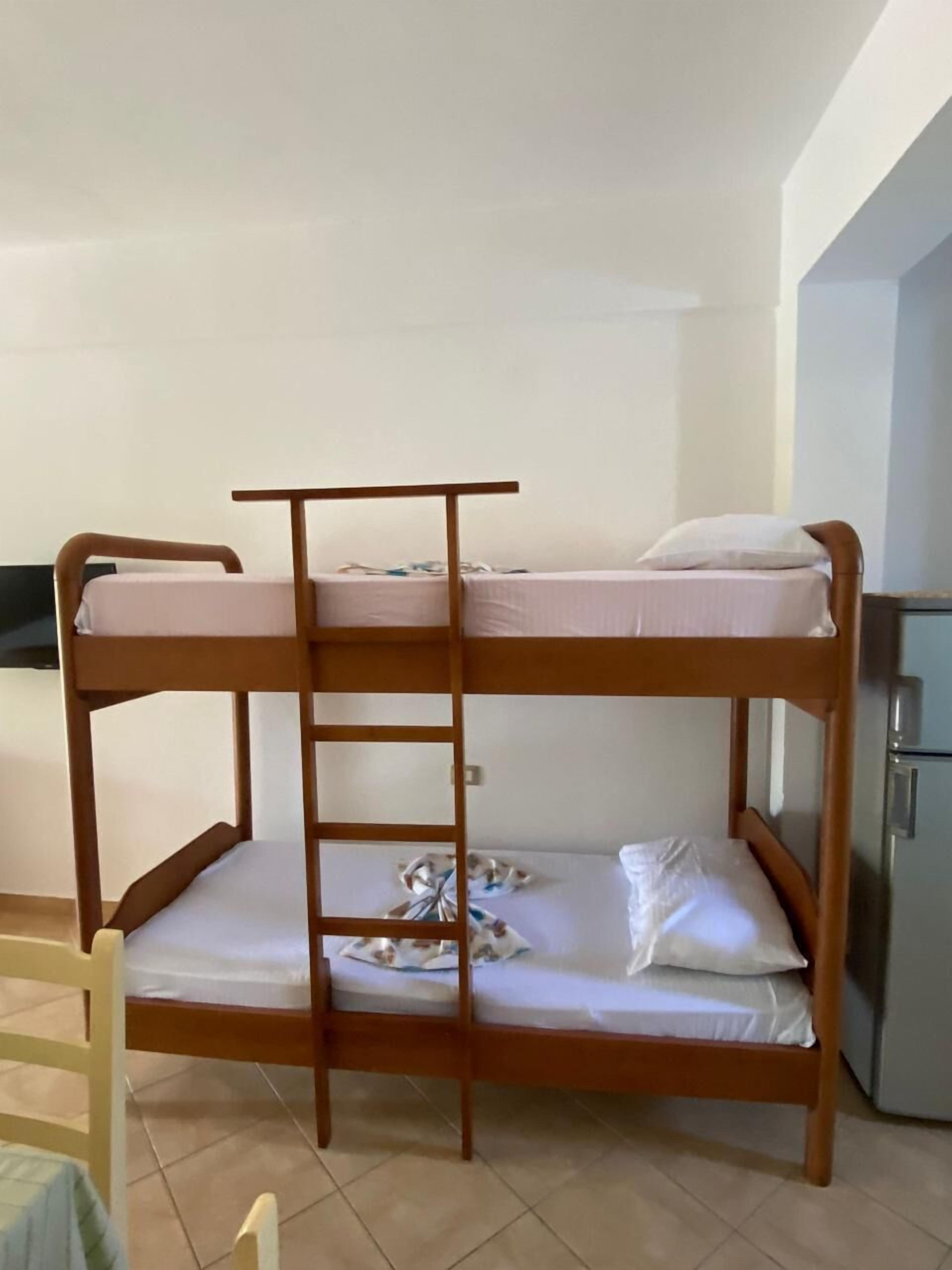A bunk bed with a ladder, a small table, and a refrigerator in a room with plain walls and tiled flooring.