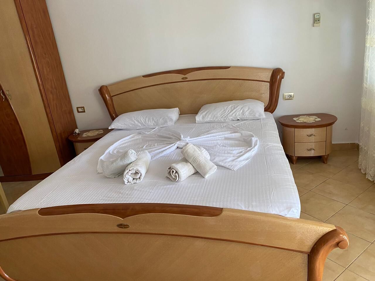 Una cama bien hecha con sábanas blancas y toallas enrolladas, un cabecero de madera, una mesita de noche con una lámpara y un armario de madera.