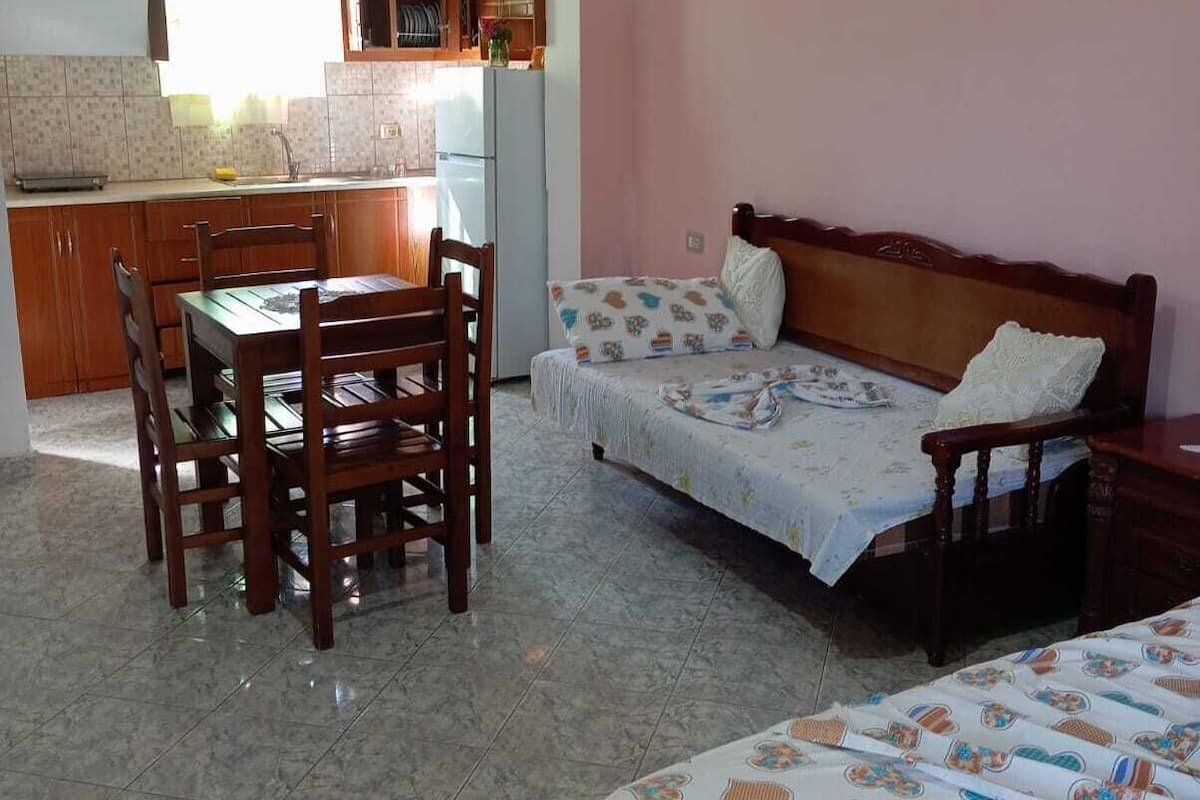 Een compacte kamer met een eettafel en stoelen, een bed met beddengoed met een patroon, en een kitchenette met een koelkast en houten kastjes.