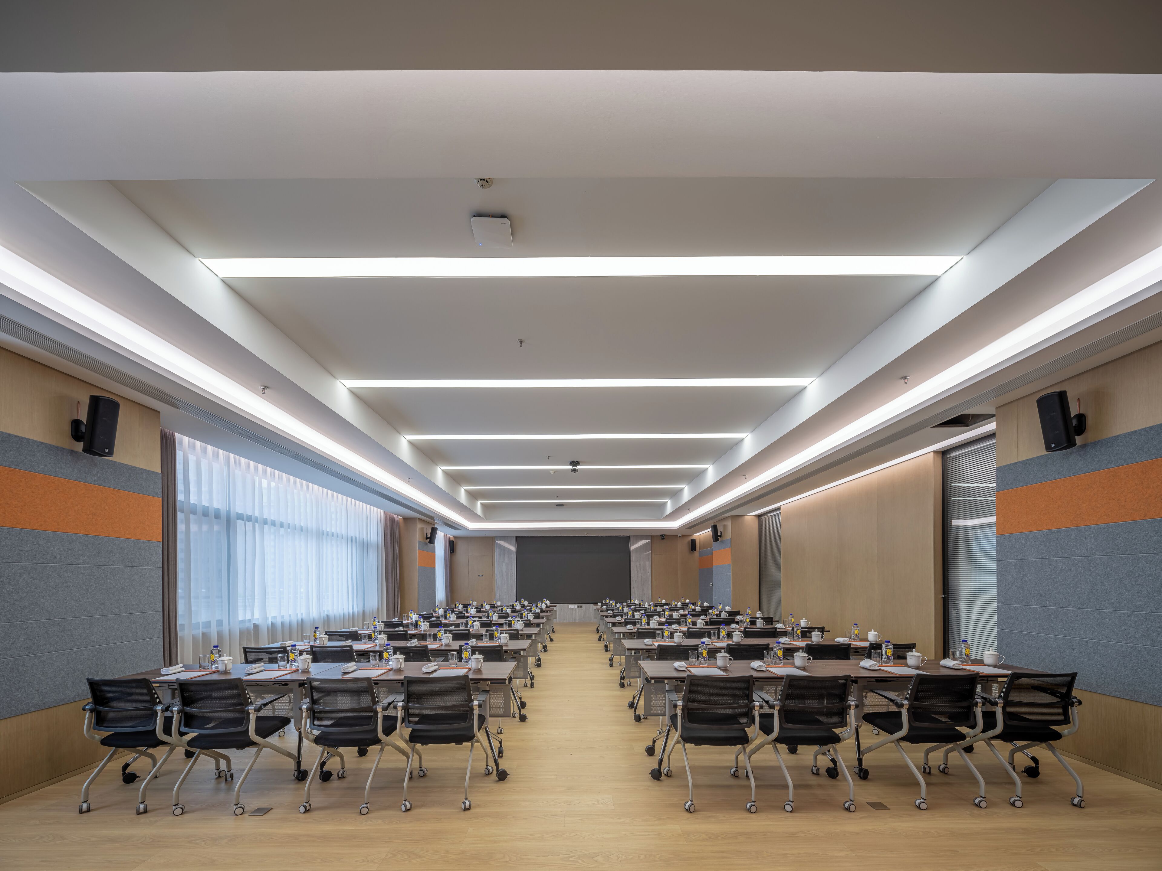 Une salle de conférence avec des rangées de tables et de chaises, des luminaires au plafond et un grand écran à l’avant.