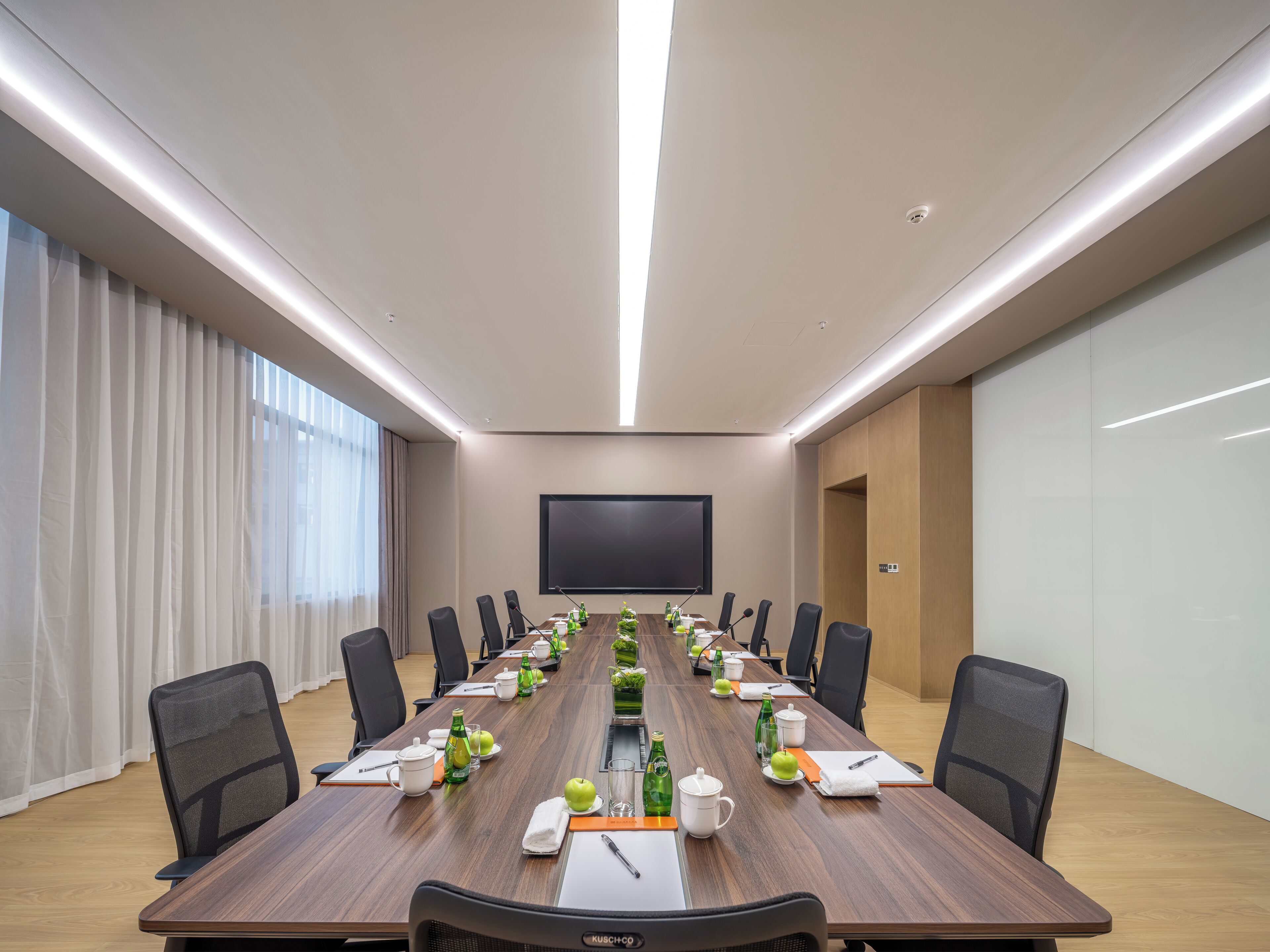 Une salle de conférence équipée d’une longue table, de chaises, d’un grand écran et de divers accessoires, comme des bouteilles d’eau et des pommes.