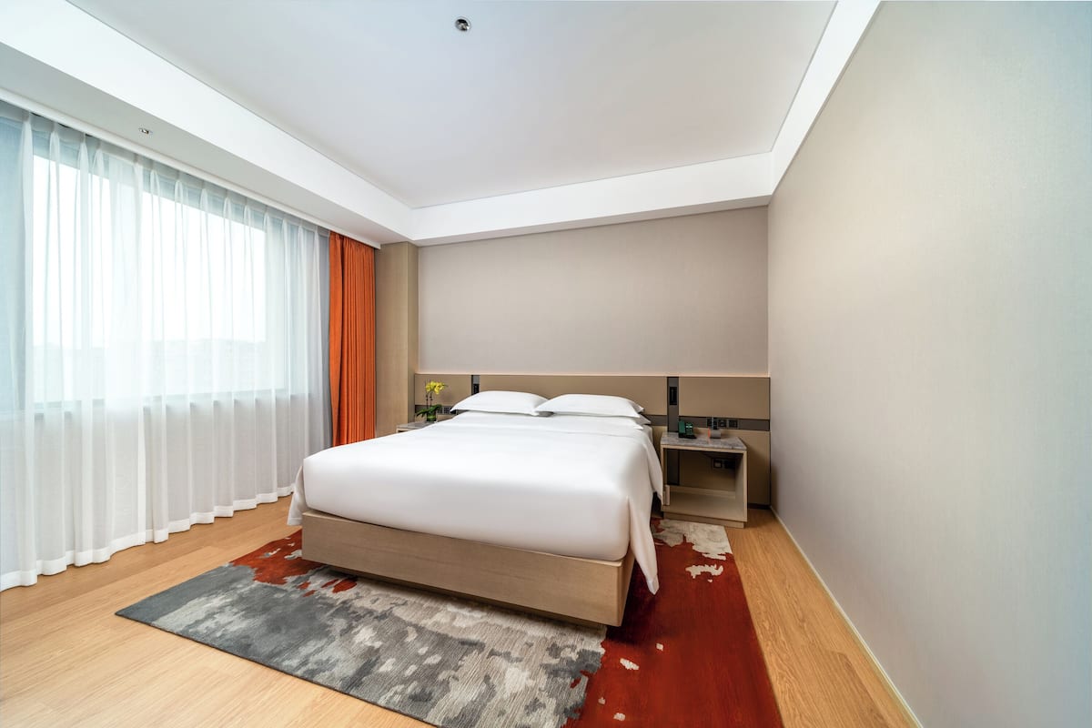 A modern hotel room with a large bed, a bedside table, a nightstand, and a window with sheer curtains.