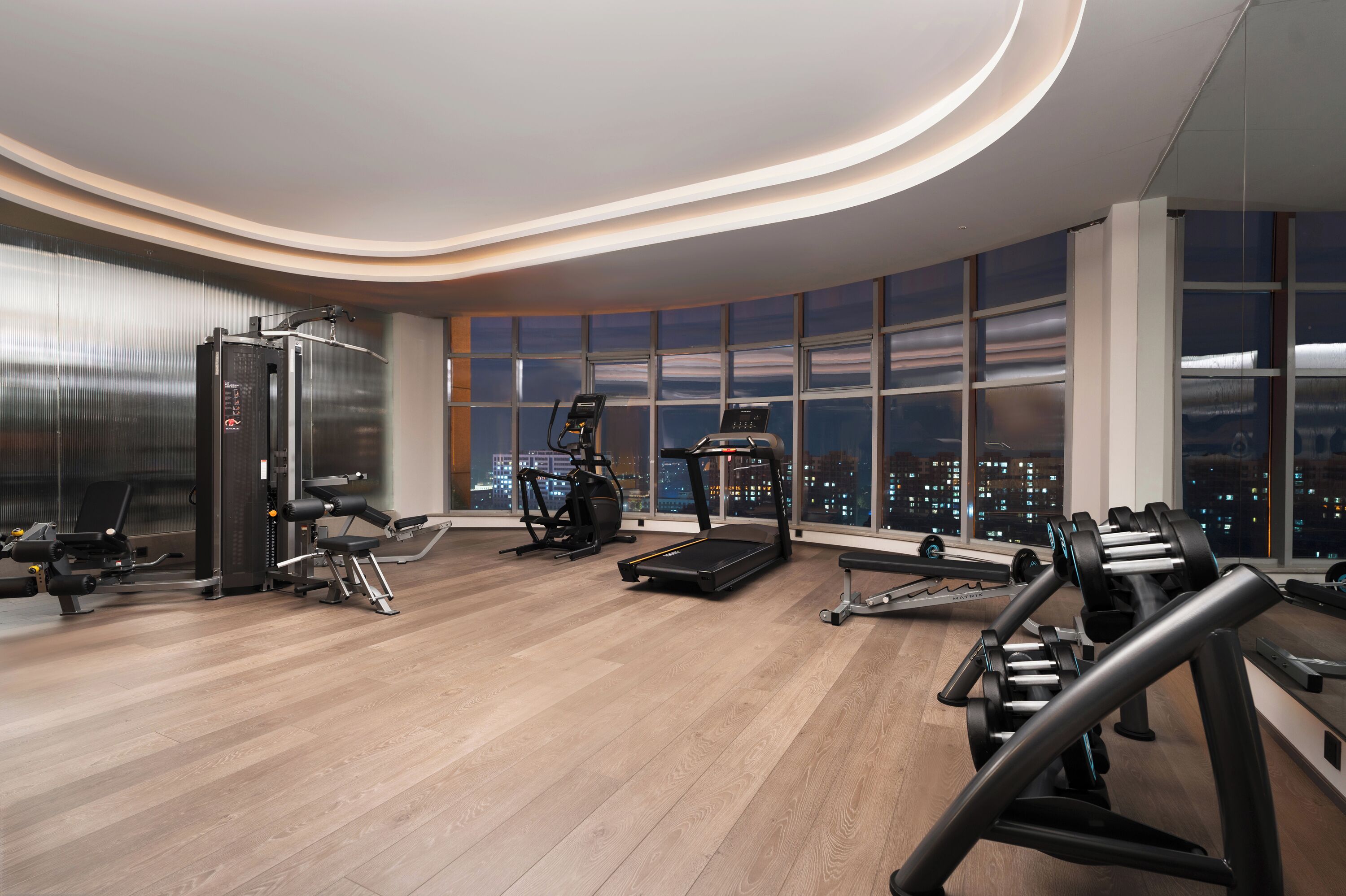 Et moderne fitnesscenter med forskellig træningsudstyr, store vinduer med udsigt over byen og et buet loftdesign.