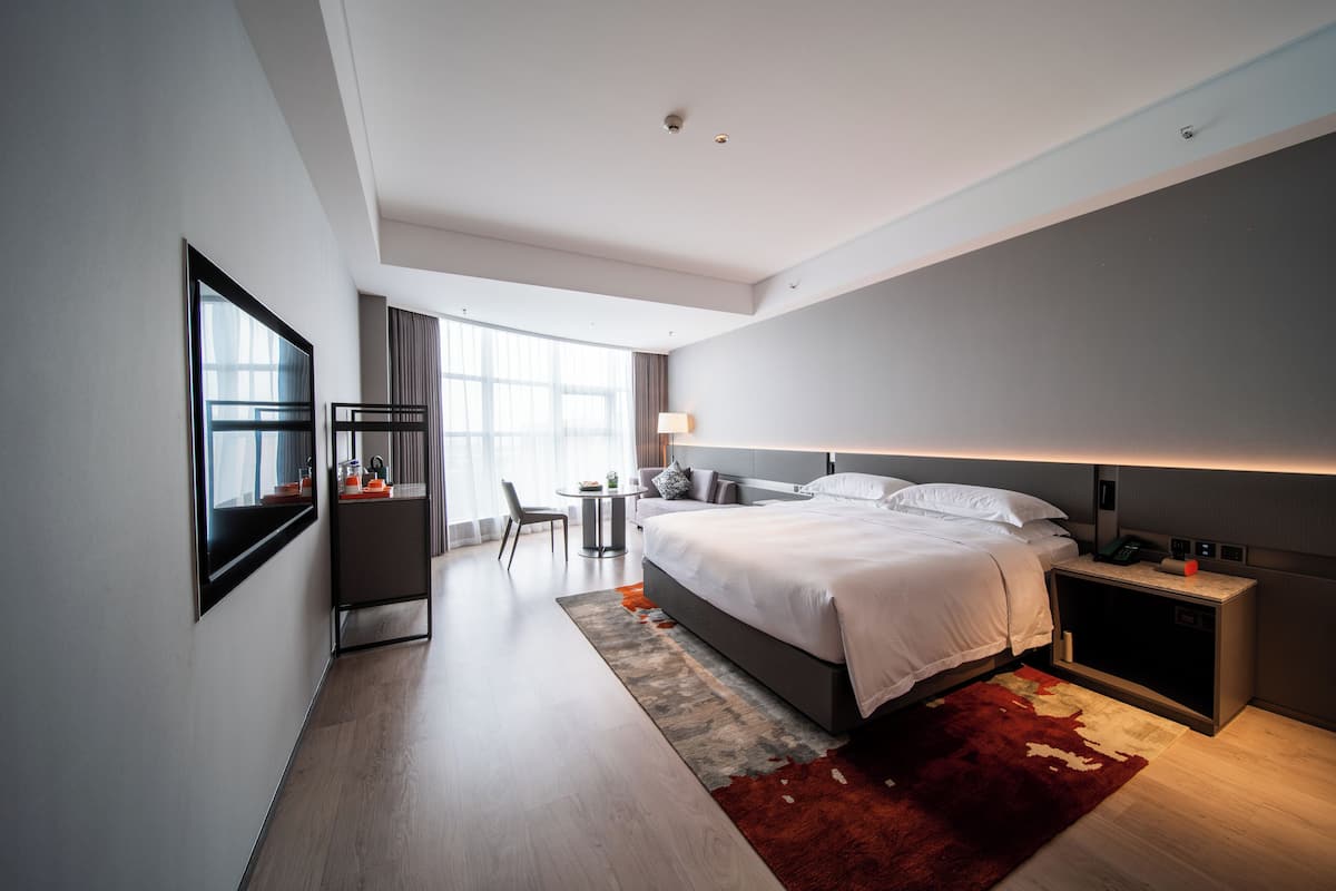 A modern hotel room with a large bed, bedside table, and a desk with a chair.