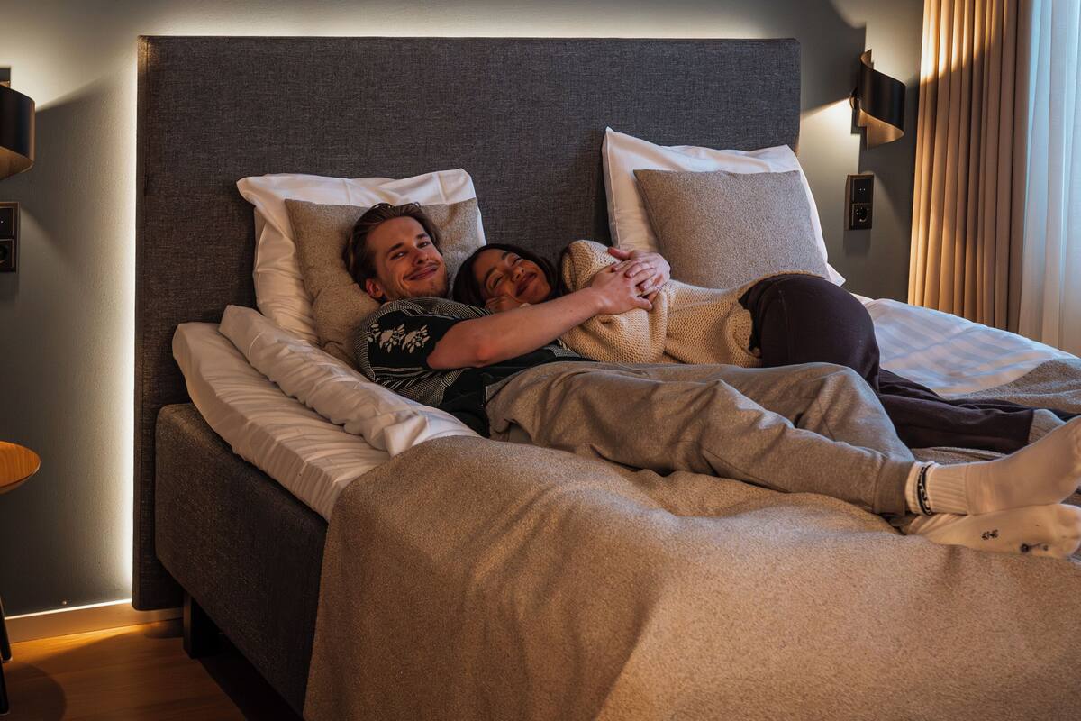A modern hotel room with a large bed, a bedside table, and a wall-mounted lamp.