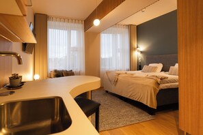 Una habitación de hotel moderna con kitchenette, una cama con cabecera y un ventanal con cortinas.