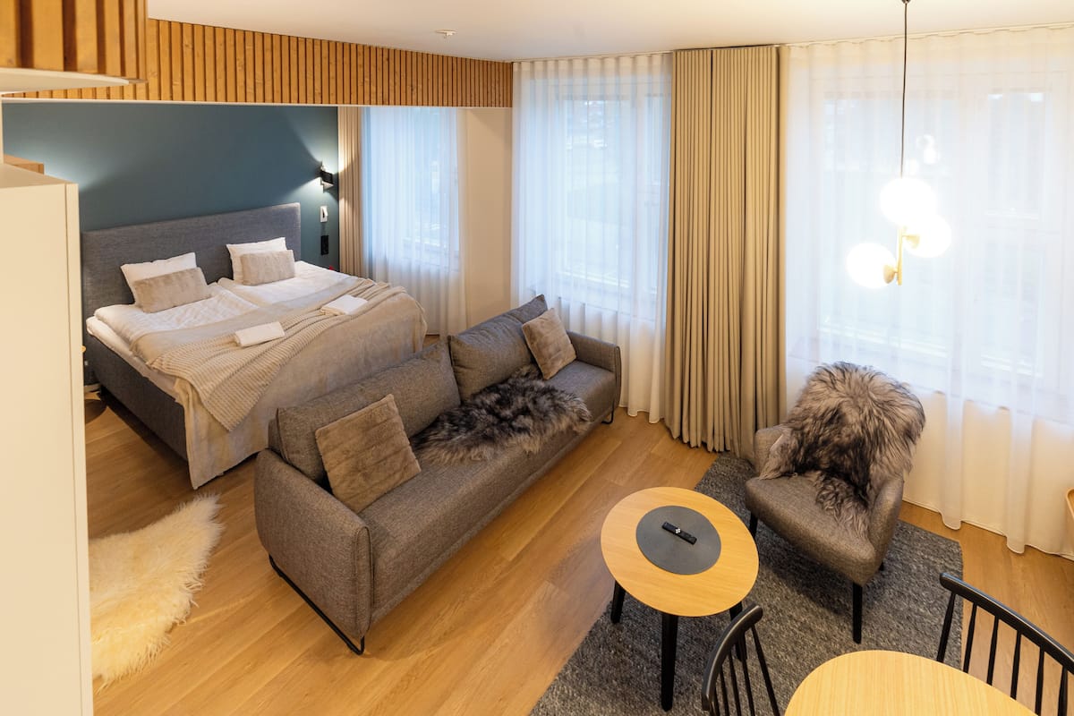 A modern hotel room with a bed, sofa, armchair, and a small table.