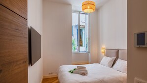 A modern bedroom with a large bed, a wall-mounted TV, a window with a view, and a ceiling light fixture.