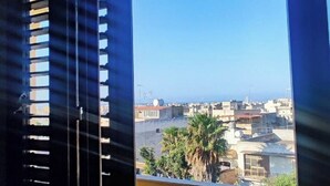 Une vue depuis une fenêtre avec des stores, donnant sur un paysage urbain avec des immeubles et des palmiers.