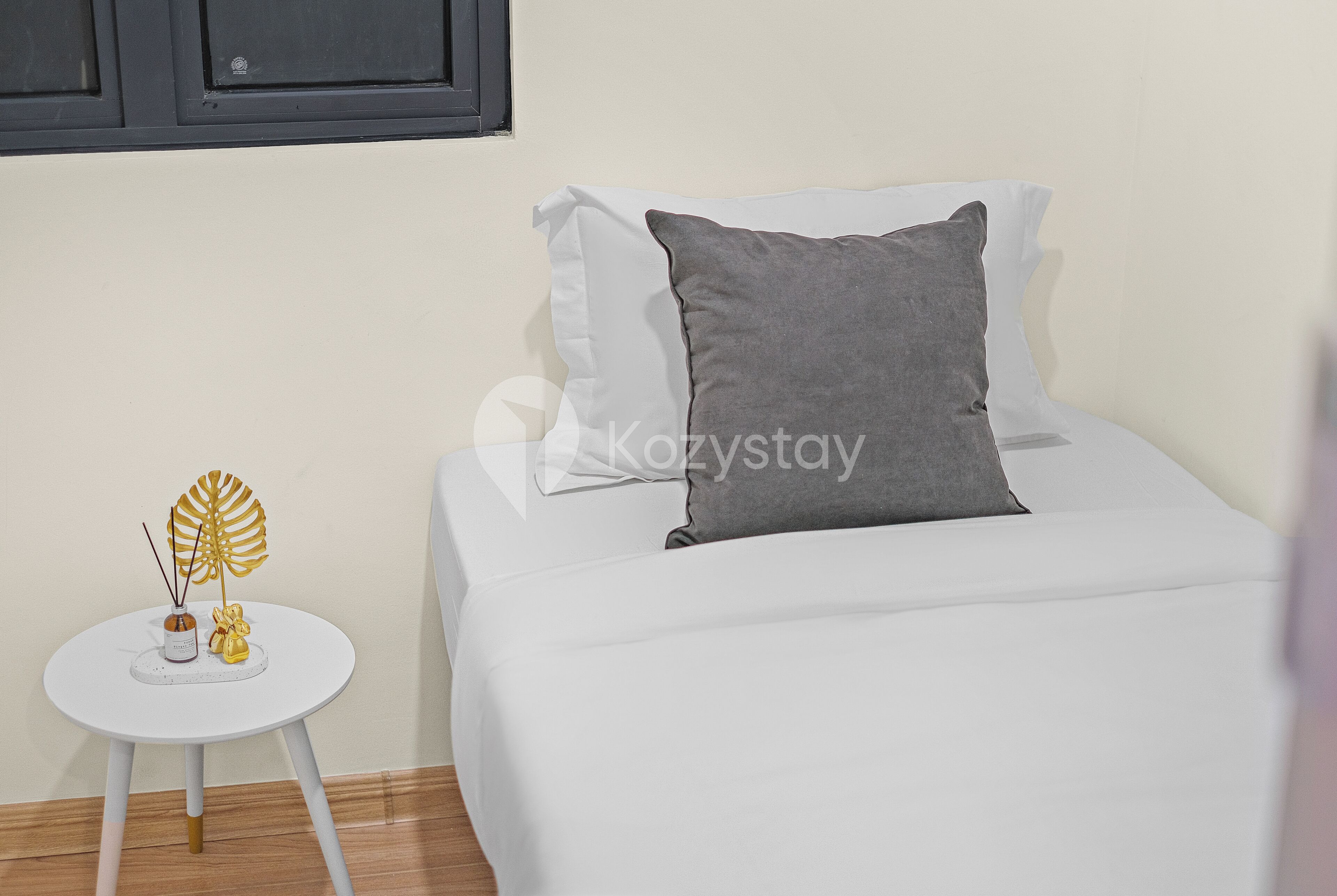 Un lit bien fait, avec un oreiller gris, une petite table blanche agrémentée d’objets décoratifs et une fenêtre avec des stores.