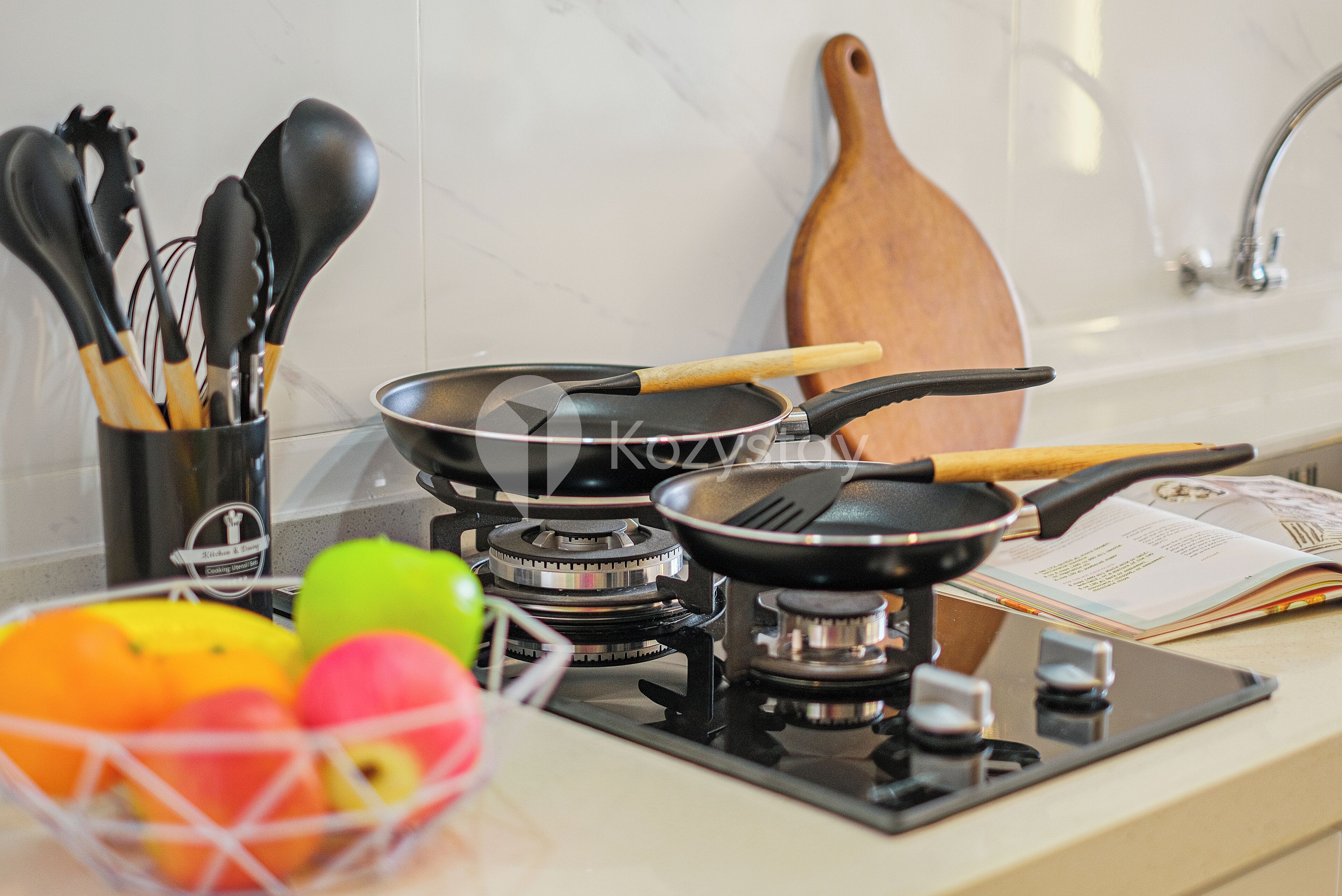 A kitchen with a gas stove, a frying pan, a spatula, and a wooden spoon. There is a bowl of fruit and a cookbook on the counter.