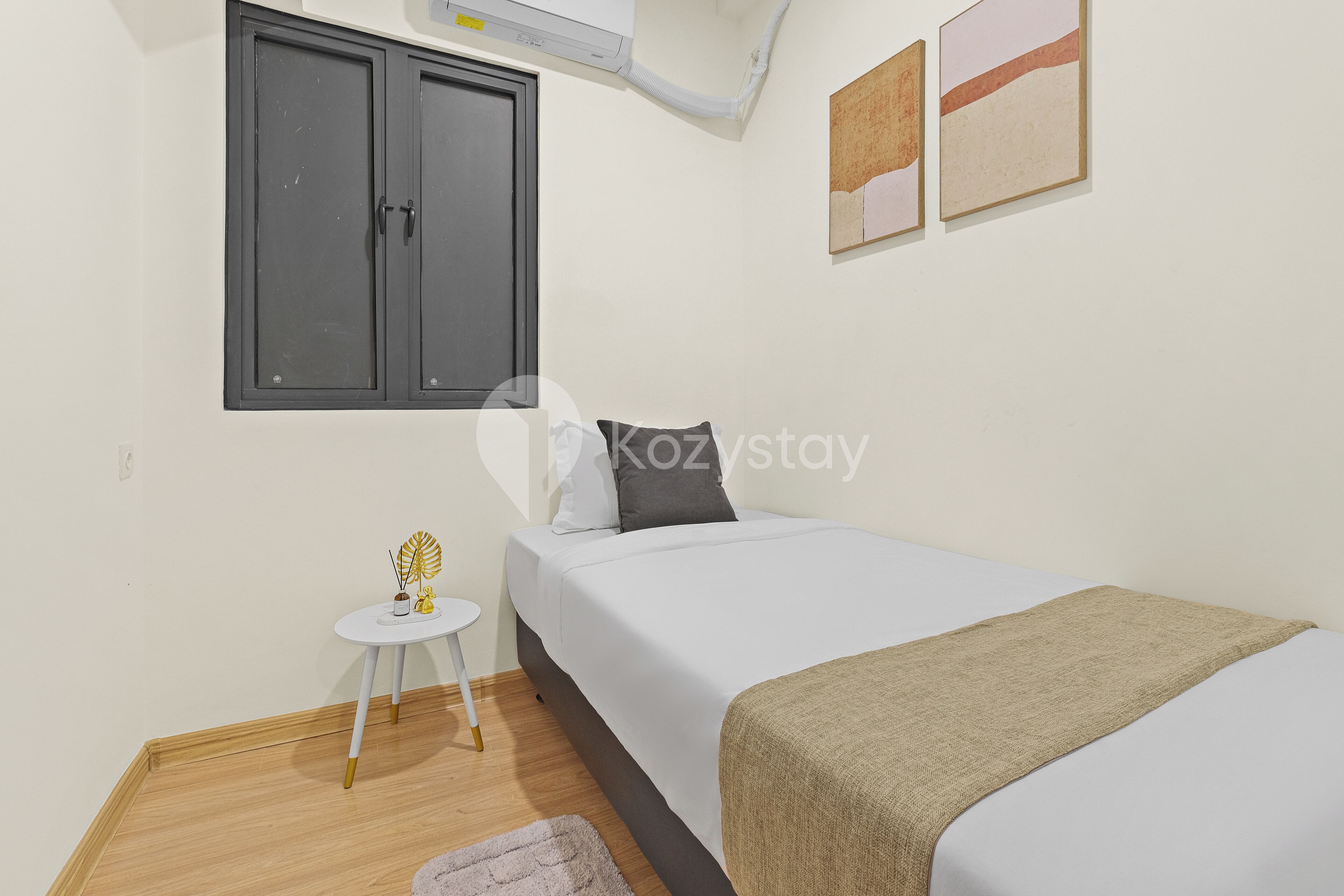 Une chambre à coucher avec un lit, une petite table avec une lampe, une fenêtre à cadre sombre et un climatiseur fixé au mur.