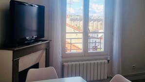 A room with a television on a wooden stand, a table with two chairs, and a window with a view of buildings and a clear sky.