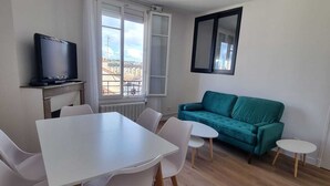 A modern living room with a dining table, chairs, a teal sofa, a television, and a window with a view.