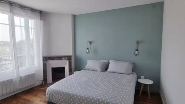 A bedroom with a bed, a small round table, a fireplace, and a window with sheer curtains.