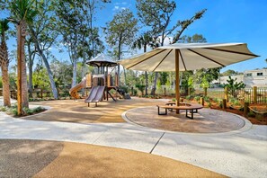 Un parque infantil con toboganes y un área de juegos, rodeado de árboles y un espacio para picnic con bancos.