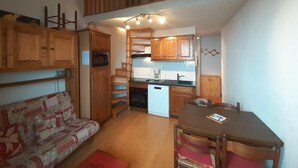 Une cuisine compacte avec des meubles en bois, un four encastré et un lave-vaisselle blanc. On y trouve une table à manger avec des chaises, ainsi qu’un canapé recouvert d’un tissu à motifs.