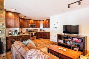 A modern kitchen with wooden cabinets, a microwave, and a stove. There is a living area with a sofa and a coffee table. A flat-screen TV is mounted on the wall.