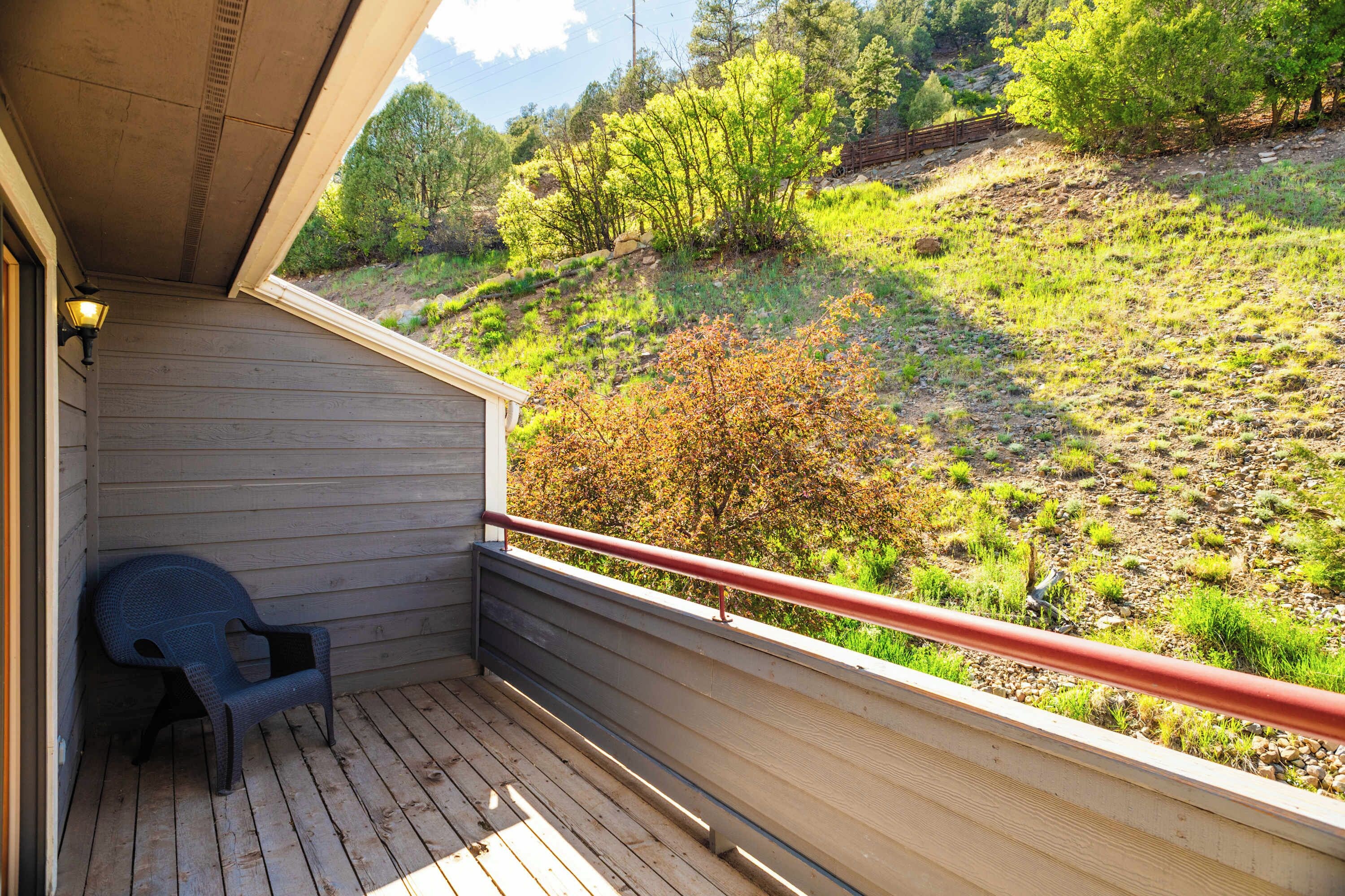 Een houten balkon met een rode reling, één stoel en uitzicht op een heuvel met bomen en struiken.