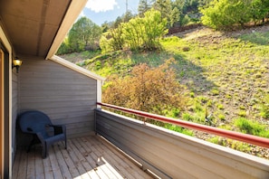 A wooden balcony with a red railing, a single chair, and a view of a hillside with trees and shrubs.