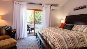 Ein Schlafzimmer mit einem großen Bett, einem hölzernen Kopfteil, einer Bank, einem Sessel und Blick ins Freie durch eine Schiebetür.