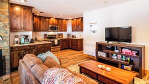 A modern kitchen with wooden cabinets, a microwave, and a stove. There is a living area with a sofa and a coffee table. A flat-screen TV is mounted on the wall.