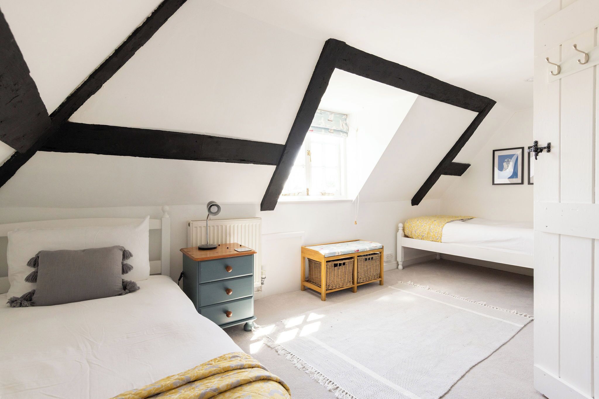 A bedroom with a sloped ceiling, a bed with a white duvet, a blue dresser, a small wooden bench, and a white door with hooks.
