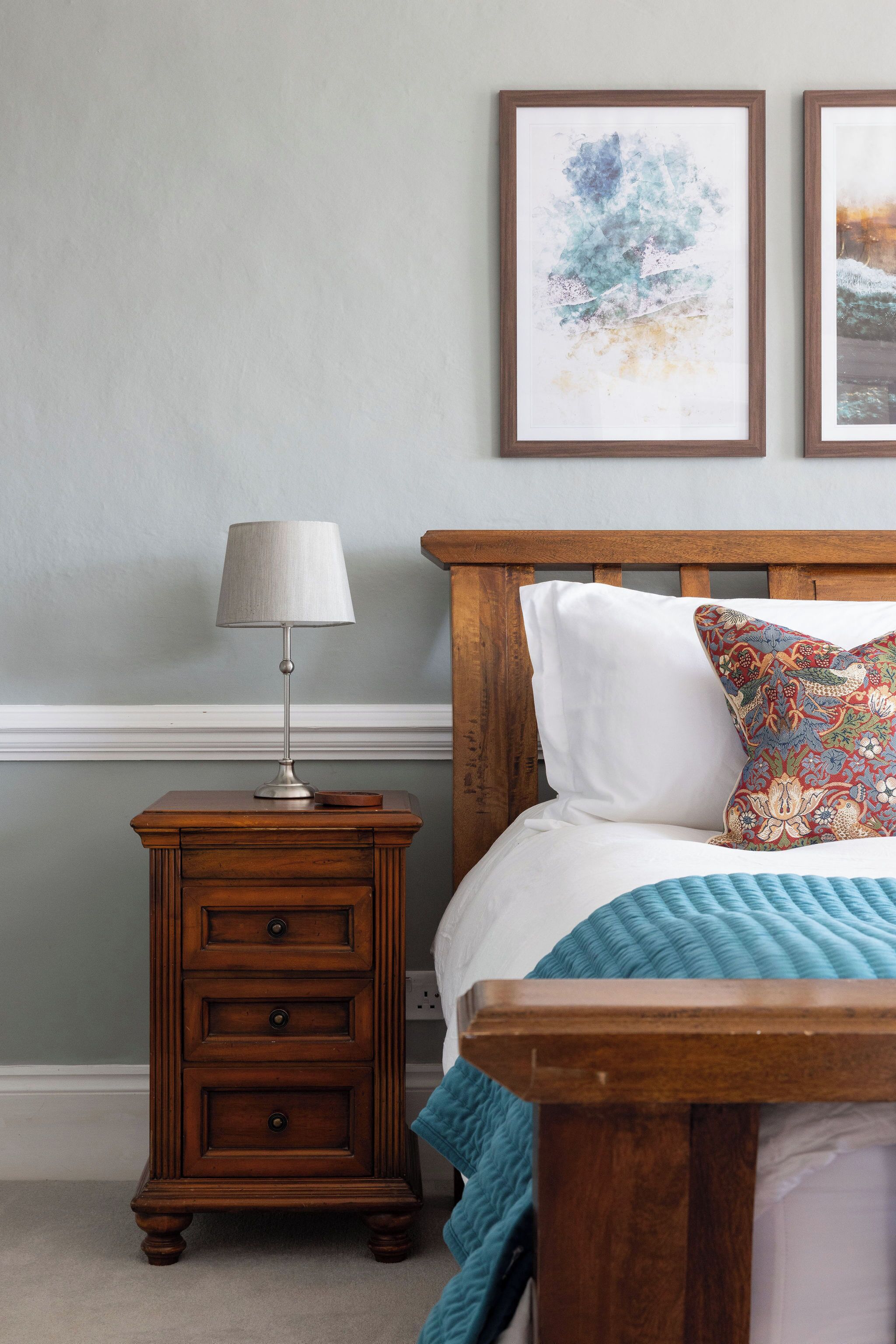 A bedroom with a wooden nightstand, a lamp, a bed with a blue quilt, and two framed artworks on the wall.