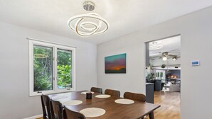 Ein Essbereich mit einem Holztisch und Stühlen, einem großen Fenster mit Blick auf Grünflächen, einer modernen Pendelleuchte und einem Gemälde an der Wand.
