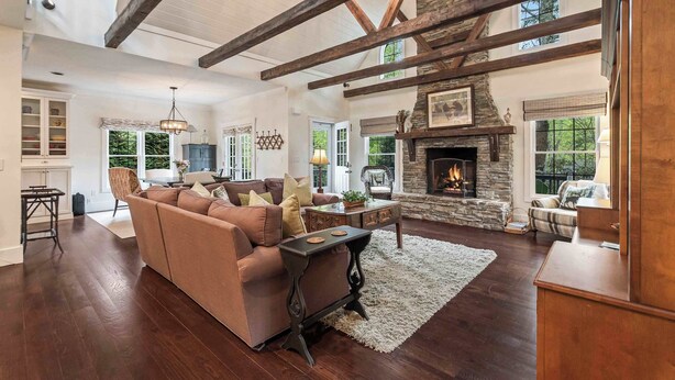 A spacious living room with a stone fireplace, wooden beams, and a large window overlooking greenery.