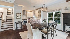 A spacious dining area with a dark wooden table and white upholstered chairs, a chandelier, and a view of the outdoors through large windows.