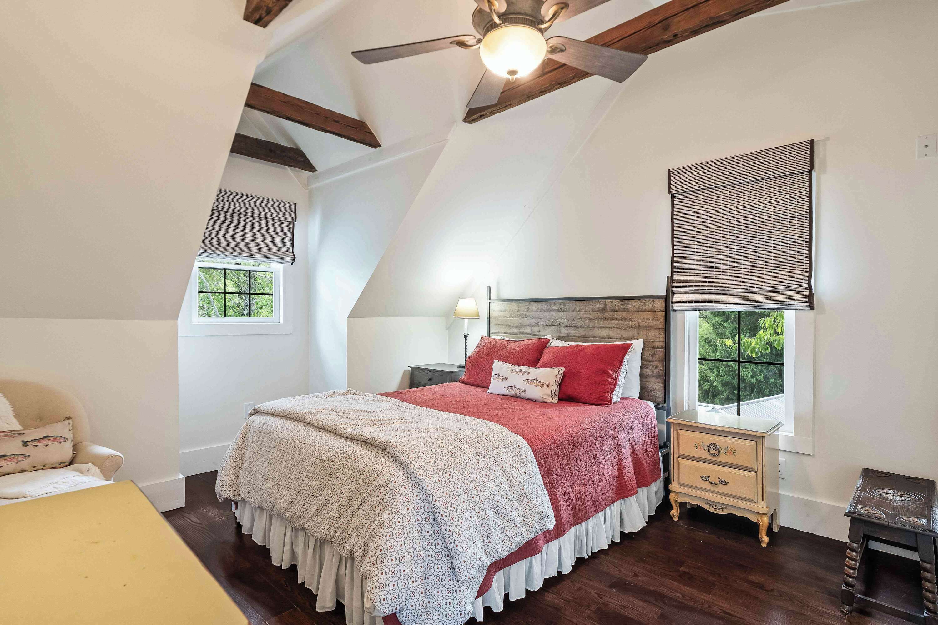 A bedroom with a sloped ceiling, a bed with a red and white bedspread, a bedside table, a nightstand, and a window with blinds.
