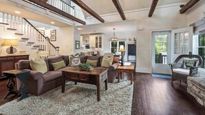 A spacious living room with a high ceiling, wooden beams, a staircase, and a view of the kitchen.