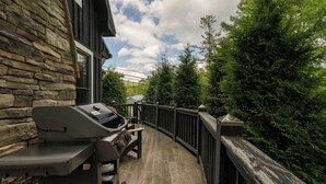A wooden deck with a barbecue grill and seating area, surrounded by trees and a stone wall.