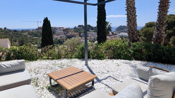 An outdoor patio with a wooden table and grey sofas, overlooking a coastal town with buildings and trees.