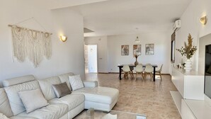 A modern living room with a beige sectional sofa, a glass coffee table, and a dining area with a white table and chairs.