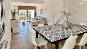 A spacious living area with a dining table, white chairs, and a sofa. There is a balcony with a view of the hills.