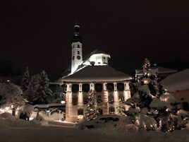Un edificio cubierto de nieve con una torre del reloj, ventanas iluminadas y árboles cargados de nieve.