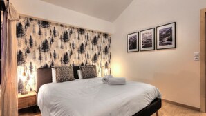 Une chambre à coucher avec un lit, une table de chevet, une lampe et un mur orné d’un motif de forêt. Trois tableaux encadrés sont accrochés au mur.