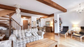 Un vaste espace de vie comprenant un canapé, des fauteuils, une table basse, des poutres en bois, un lustre et un escalier.