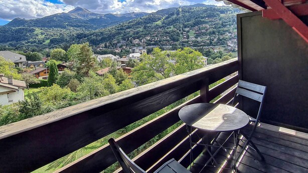 Un balcon avec une table et des chaises, donnant sur un paysage montagneux avec un village en contrebas.