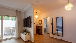A modern living room with a flat-screen TV, wooden flooring, a large mirror, and a ceiling light fixture. - The Secret Garden Stay (Agia Paraskevi)