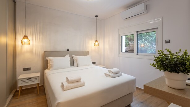 A modern hotel room with a large bed, bedside tables, a potted plant, and a window with a view of greenery. - The Secret Garden Stay (Agia Paraskevi)