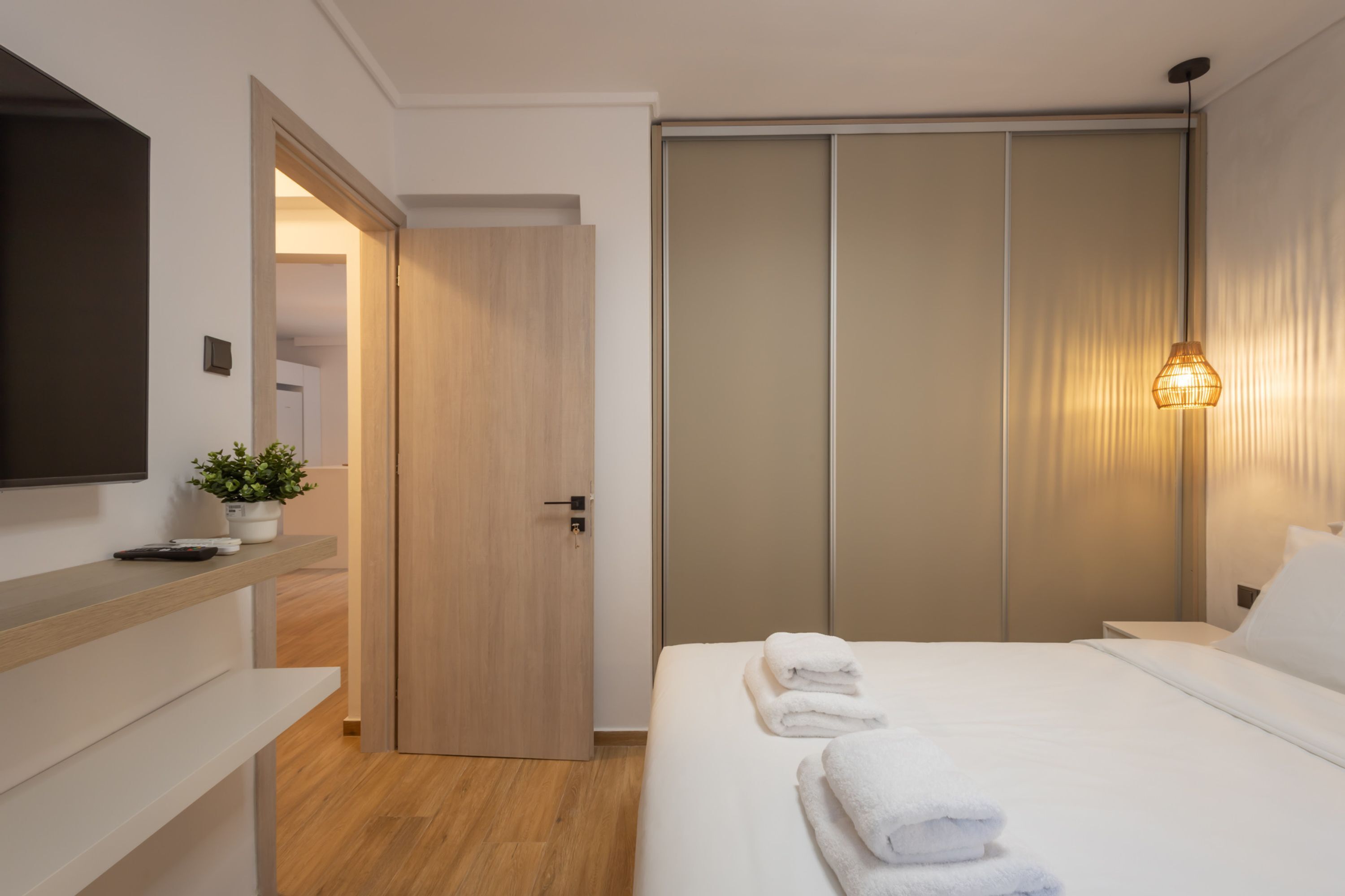 A modern hotel room with a wooden door, a bed with white linens, a bedside table with a plant, and a wall-mounted TV.