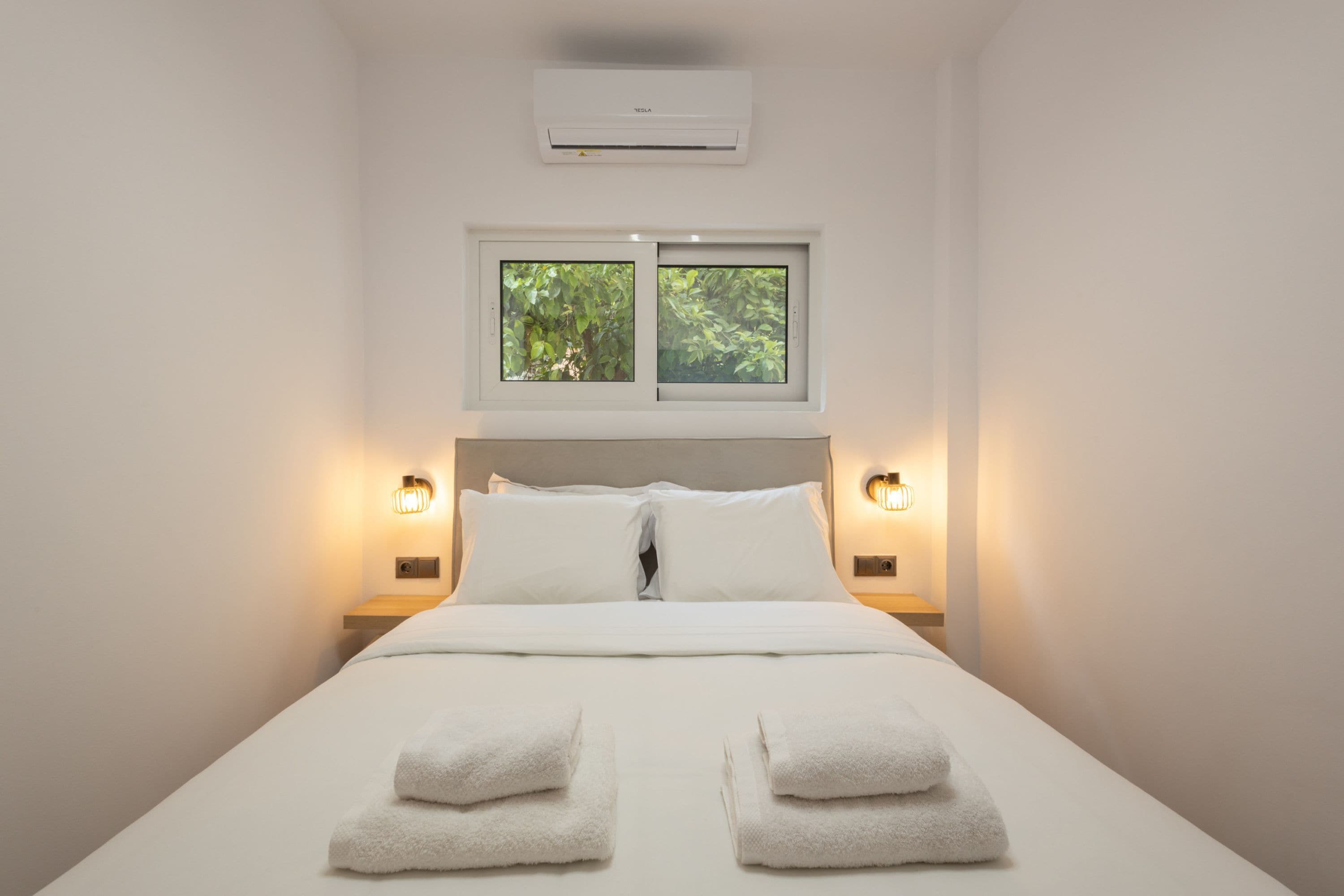 A hotel room with a bed, two windows, and wall-mounted air conditioning.