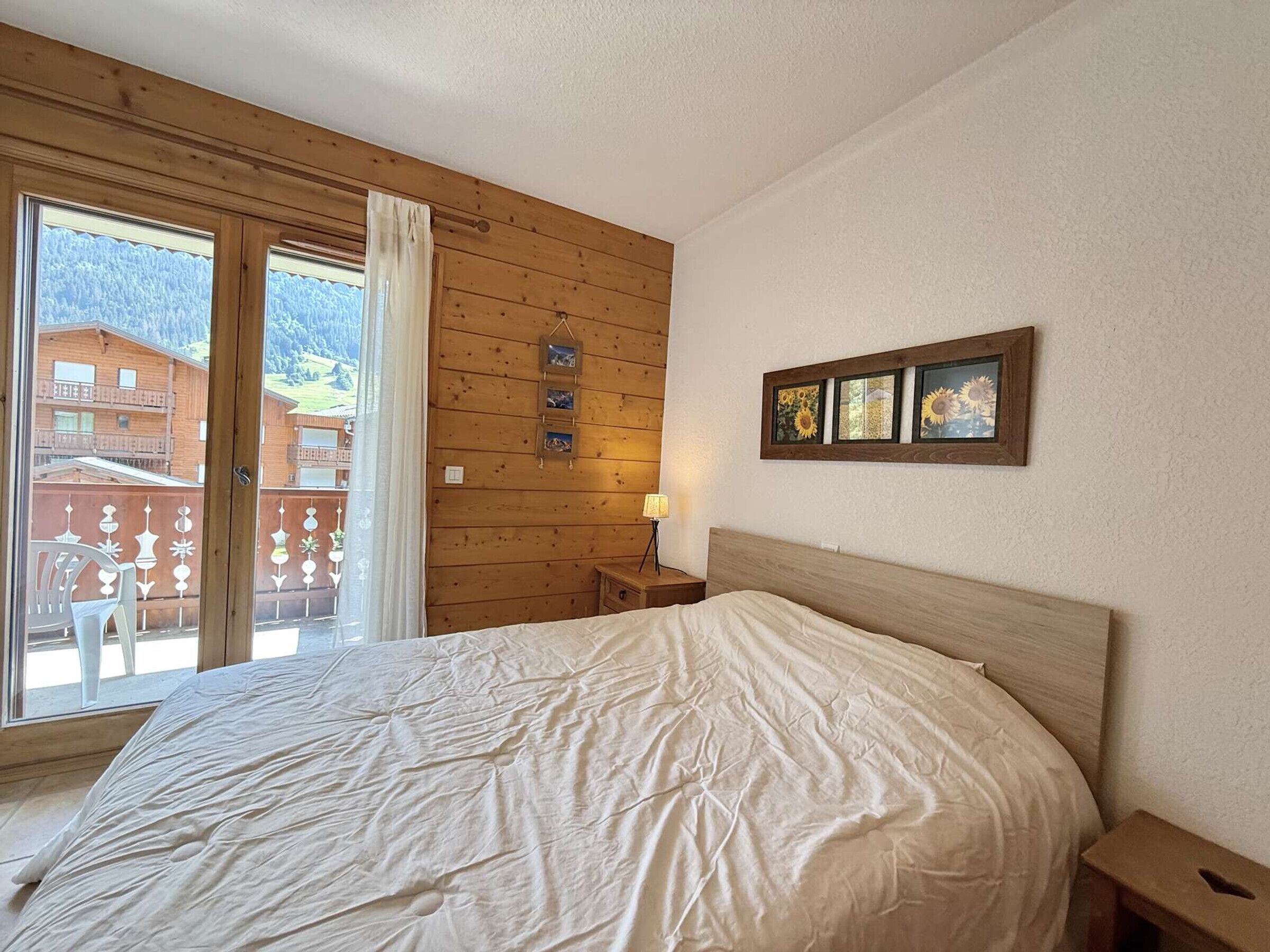 A bedroom with a large bed, wooden walls, a window with a view of buildings, and a small bedside table with a lamp.
