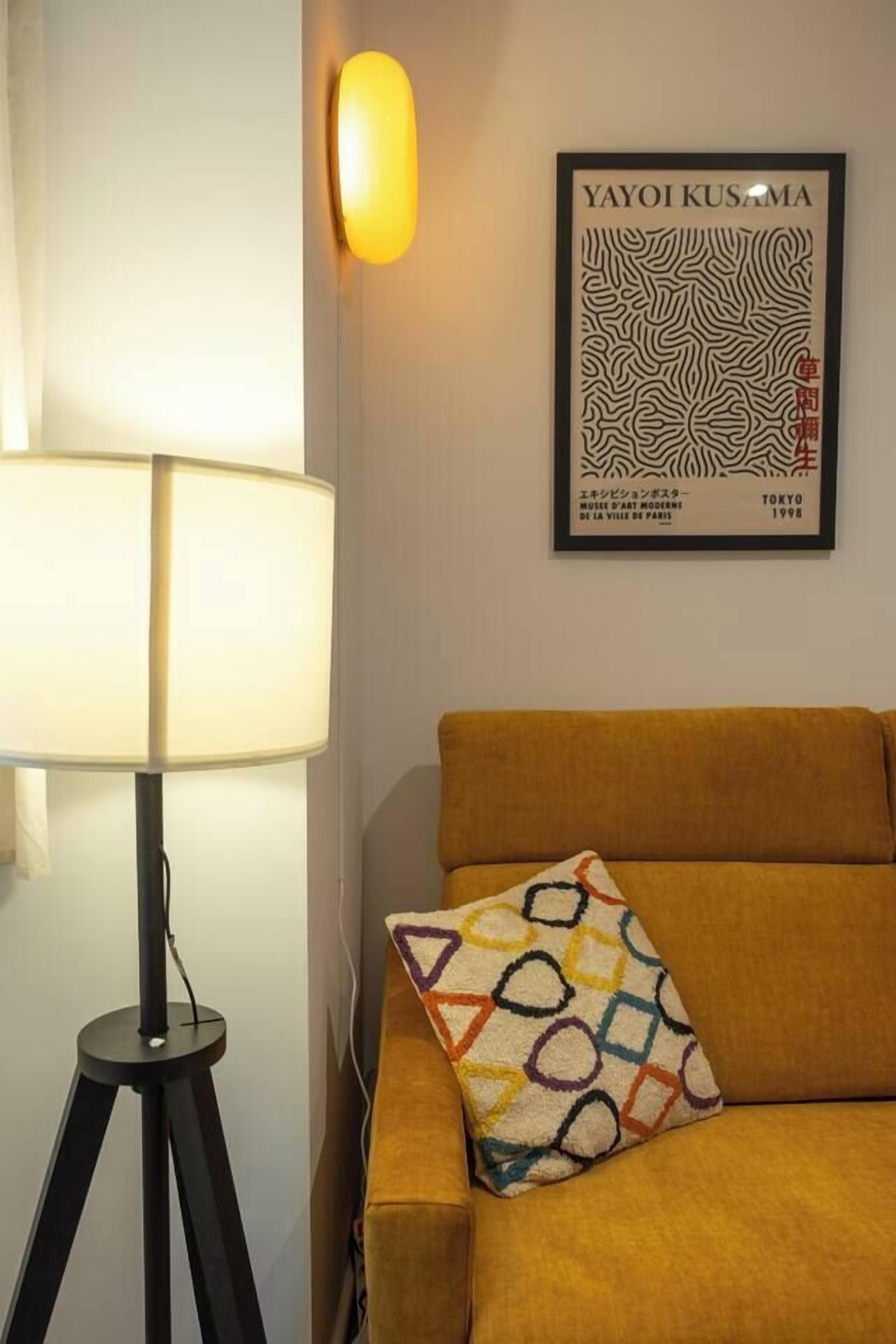 A room with a lamp on a tripod, a sofa with a patterned cushion, and a framed artwork on the wall.