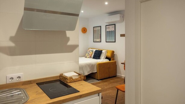 A compact hotel room with a bed, a small table, a built-in stove, and a wall-mounted air conditioner.